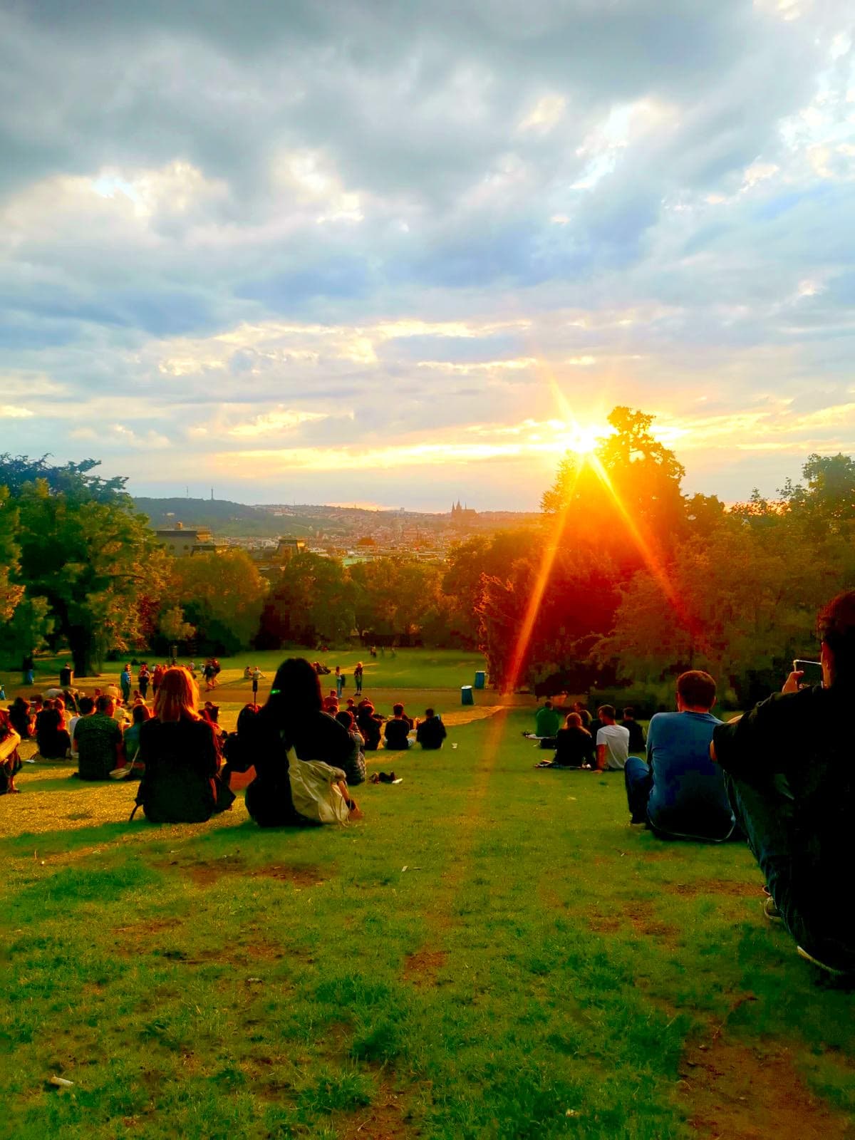 A crowd of people sitting in Riegrovy Sady Park watching the sunset over Prague's Old Town with Prague Castle in the distance.