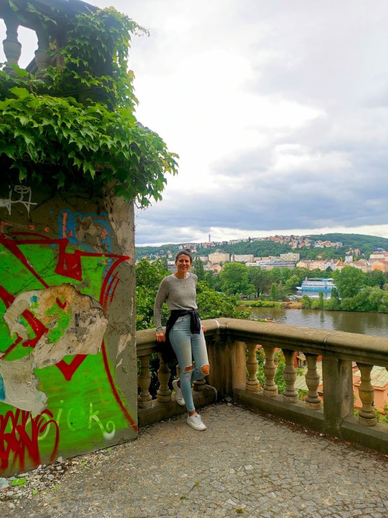 Katharina standing in front of a stone fence on the way up to Vyšehrad Fortress with a green hill and Vltava River in the background.