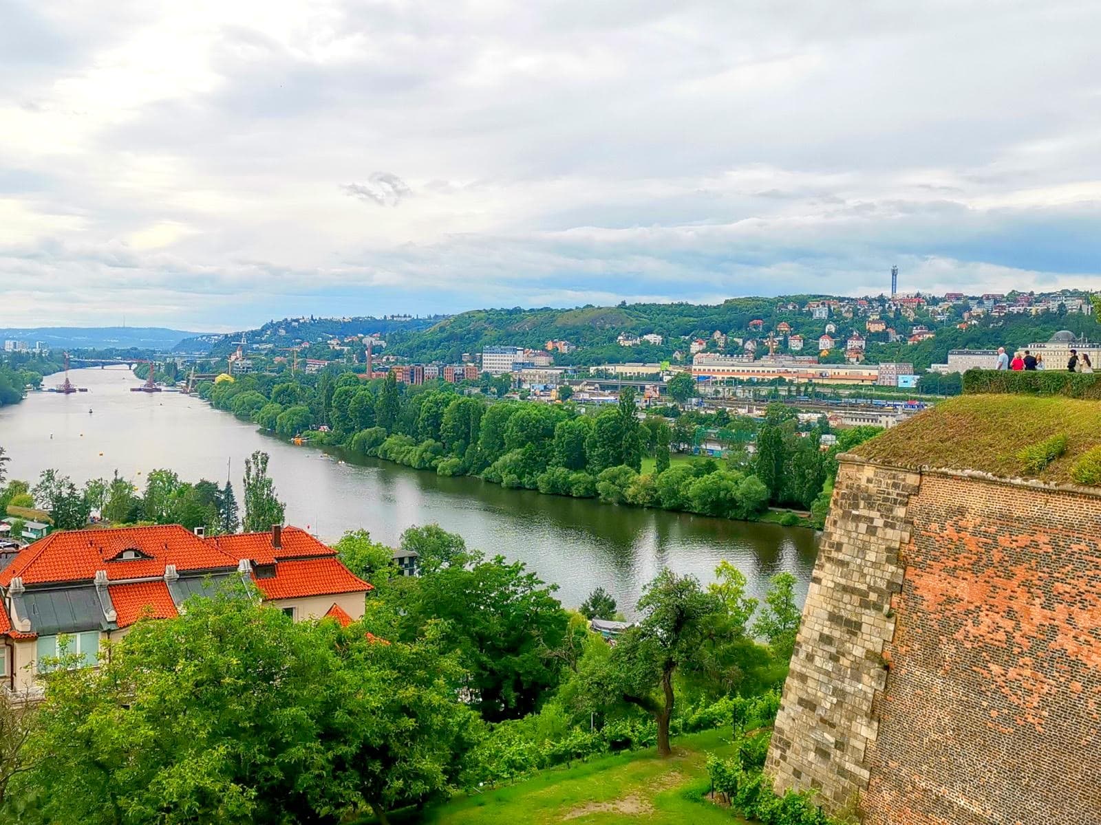 Scenic view over the Vltava River, with lots of trees and distant houses, alongside a red brick wall of Vyšehrad Fortress, a historic landmark to see on a 2-day Prague itinerary.