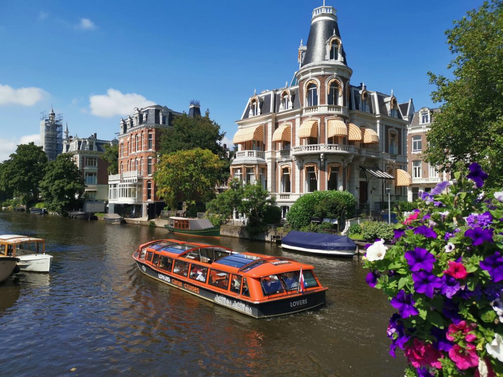 A orange boat cruising down one of the rivers in Amsterdam with colorful flowers in the foreground and a beautiful old building in the background