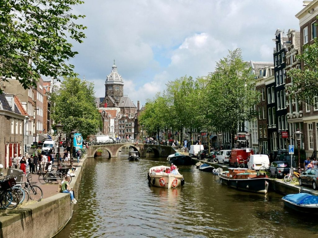 A tour boat cruising down a canal in amsterdam while a couple of people sit on the edge of the canal with their legs dangling over the edge