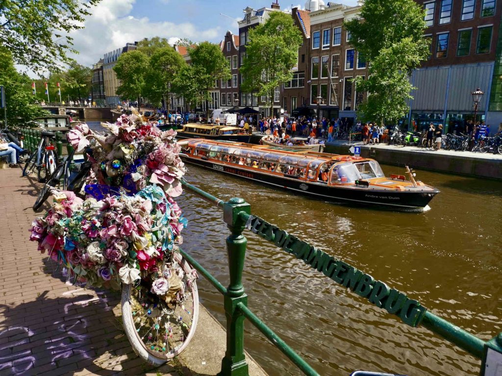 A bike covered in flowers leaning next to a railing by a canal in amsterdam with a boat full of tourists going on a boat cruise in the background