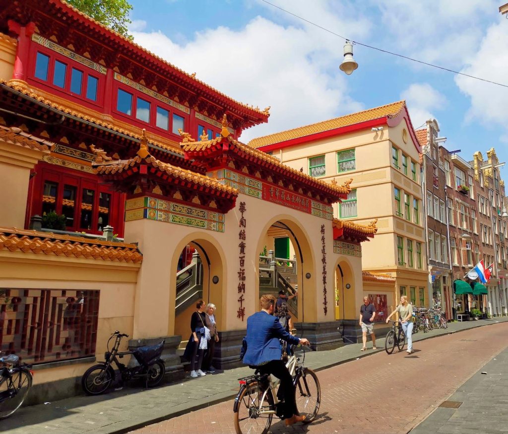 A man in a suit riding his bike down the street next to a chinese temple in the middle of Amsterdam