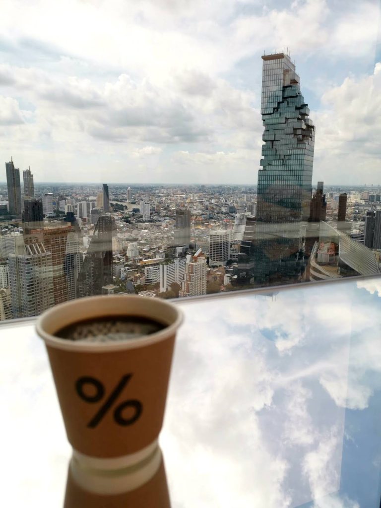 A paper cup with a %-symbol, filled with black coffee, placed in front of a large glass window overlooking the iconic King Power Mahanakhon Tower in the distance.