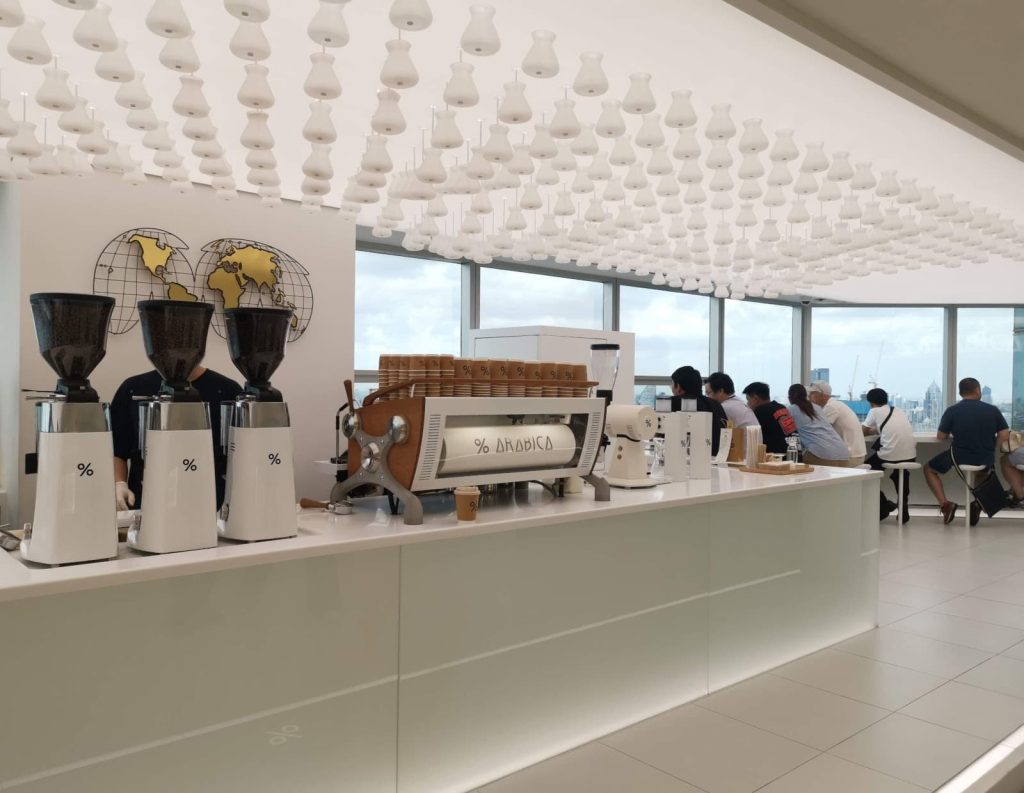 Interior of % Arabica Bangkok Empire Tower Cafe featuring a large white coffee bar at the center, a world map on the wall, and people seated by large windows overlooking the city view.