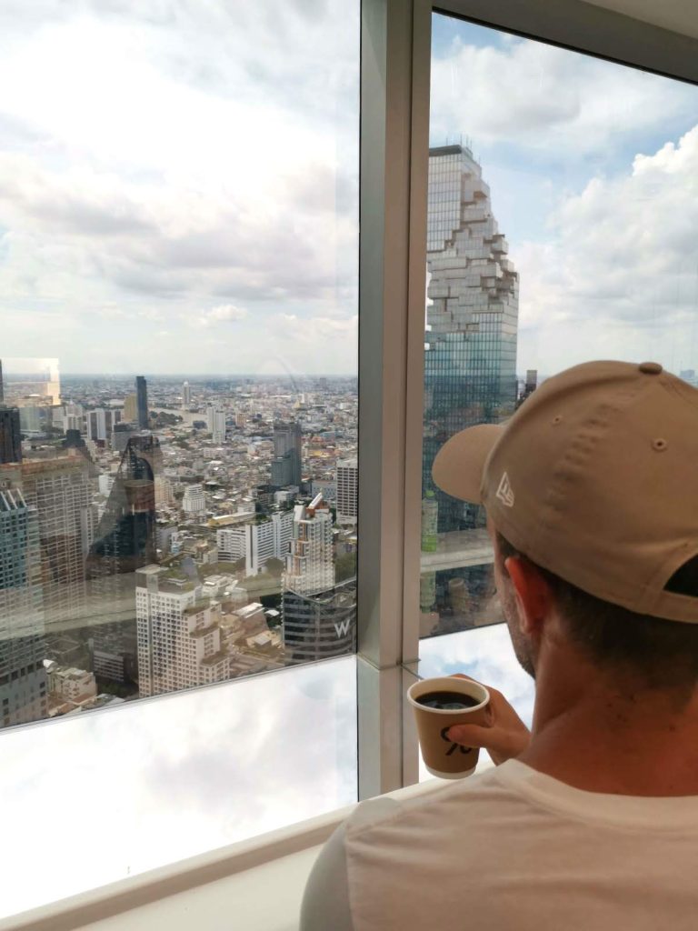 Allan holding a paper coffee cup, looking out the window at a view of Bangkok's skyscrapers.