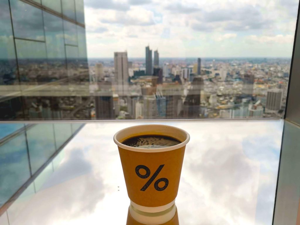 Paper cup with black coffee in front of a large window overlooking a city skyline with skyscrapers, at one of the best cafes in Bangkok with a view.