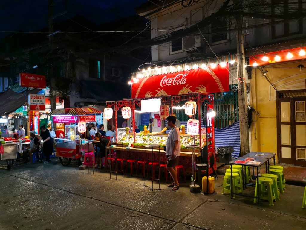 People walking down a street in Bangkok's Chinatown at night with various different street vendors selling food.