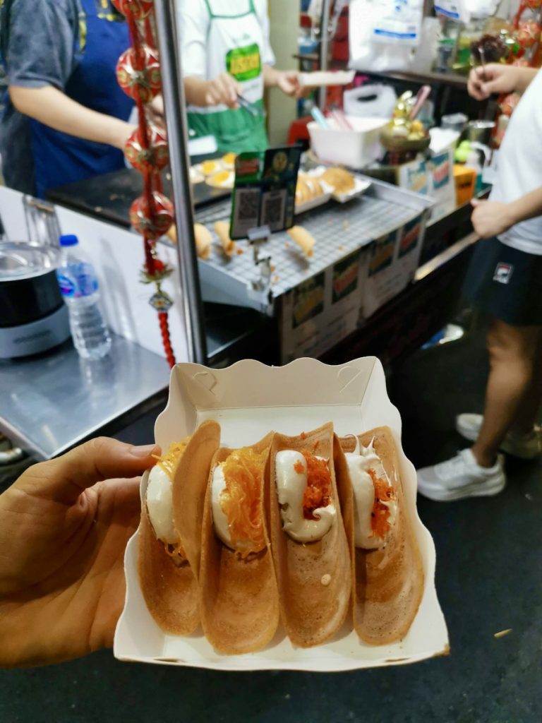 My hand holding a small white cartboard plate with a serving of four crispy coconut pancakes on top.