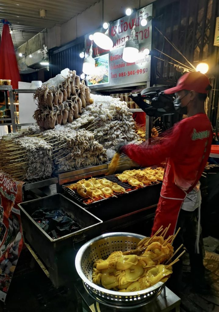 A Chinatown Bangkok street food stall with a huge pile of squid sitting next to a grill and a cook preparing it.