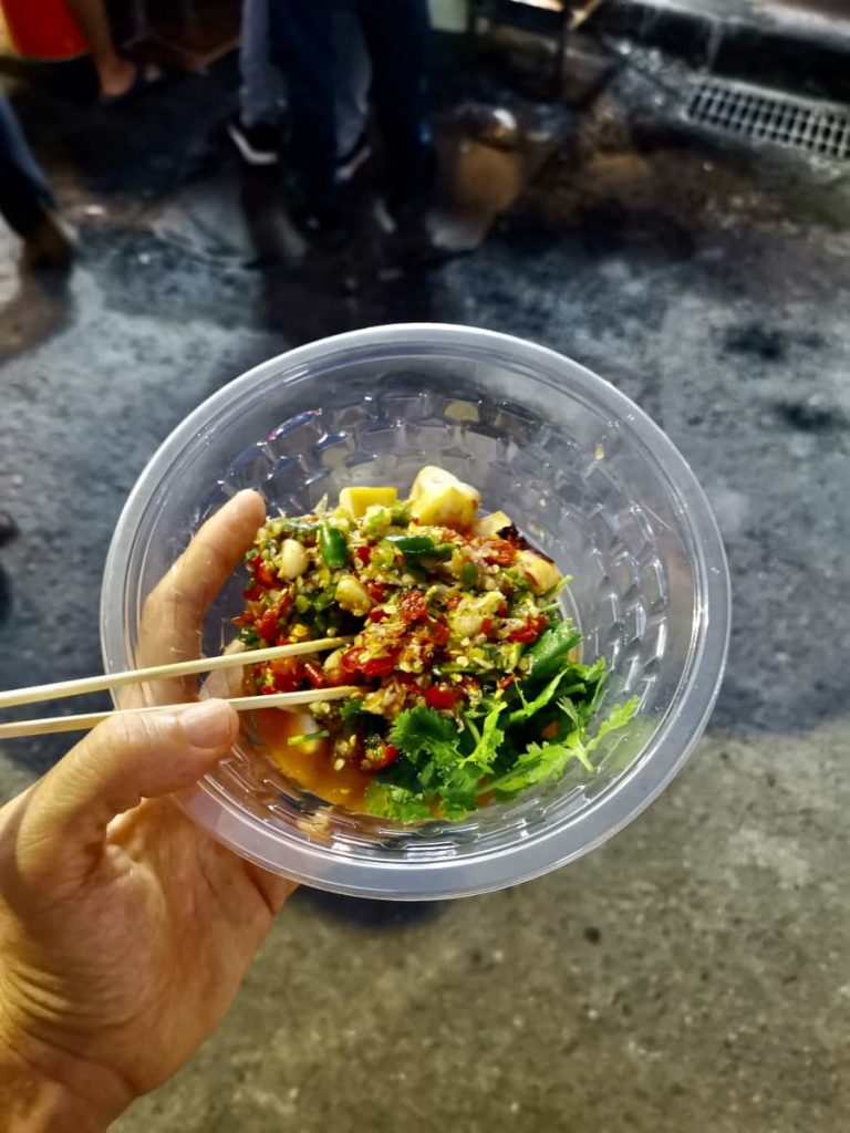 My hand holding a plastic bowl with wooden chop sticks that has pieces of grilled squid in a spicy chilly sauce and lettuce in it.