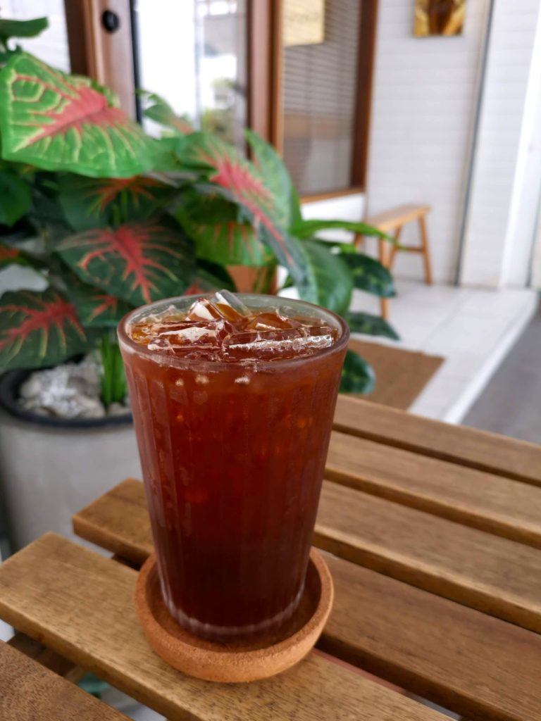 Glass of black coffee with ice cubes on a wooden table, with a green plant in the background.