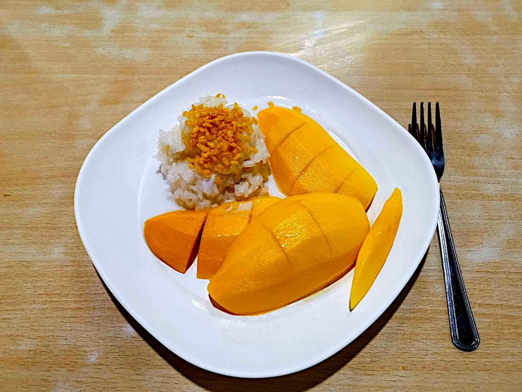 A white plate on a wooden table with cut pieces of yellow Mango and a portion of white rice with crispy mung beans on top next to it.