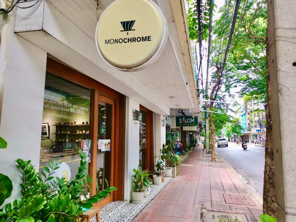 Wooden door entrance of Monochrome Cafe on a quiet, green leafy street in Bangkok's Silom district.