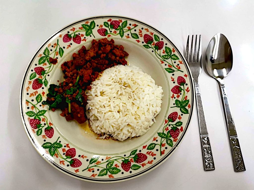 A plate of Pad Kra Pao, a popular dish in Thai cuisine, on a plate on a table with a fork and spoon next to it.