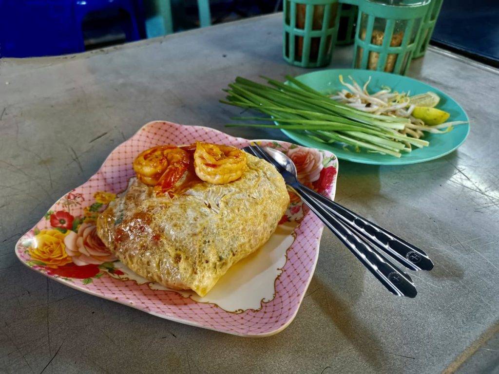 A pink plate with a serving of Pad Thai on it that is wrapped inside a layer of egg with two shrimp on top of it.