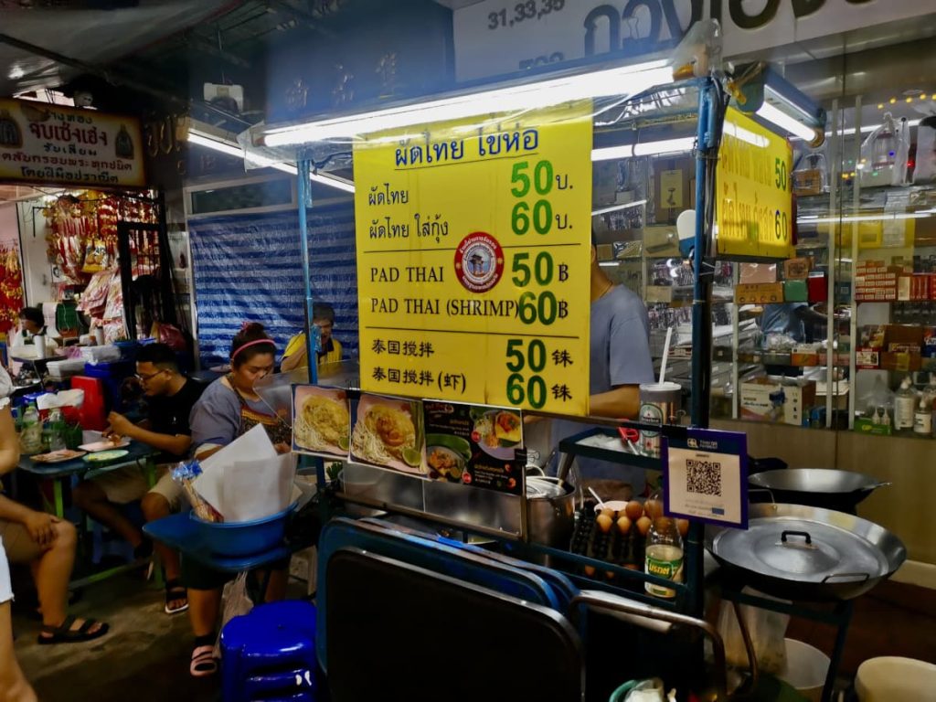 A small food cart in Chinatown Bangkok selling Pad Thai with a yellow sign on it displaying the menu options and prices in Thai Bhat.