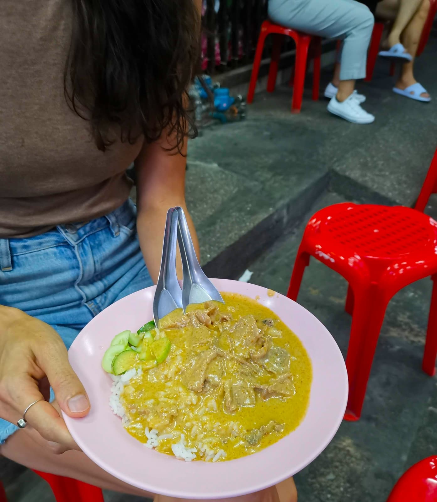 A bowl of red Thai curry with two spoons in it sitting on the knees of Katharina at a Netflix-famous Chinatown Bangkok street food stall.