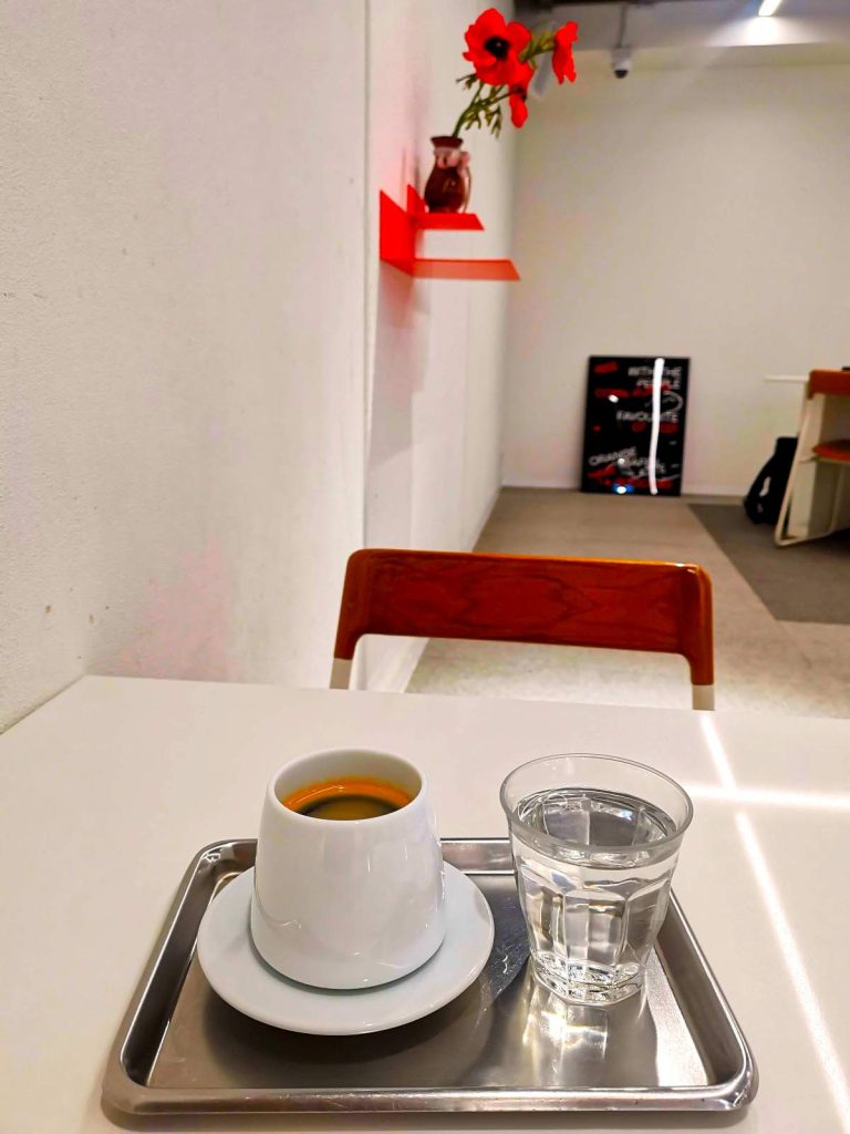 Black coffee in a white cup beside a glass of water on a white table, with orange flowers in the background.