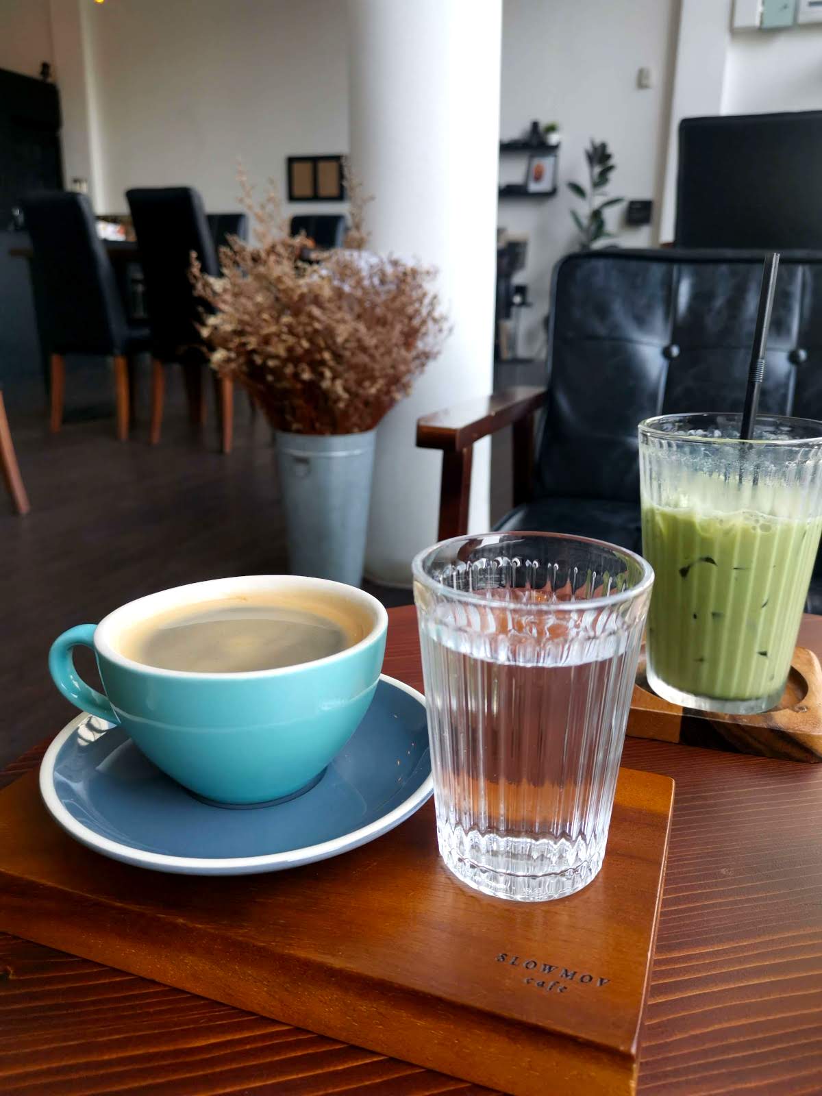 Hot Americano in a light blue ceramic cup, next to a glass of water and a green Matcha Latte on a wooden table, with black leather seats in the background.