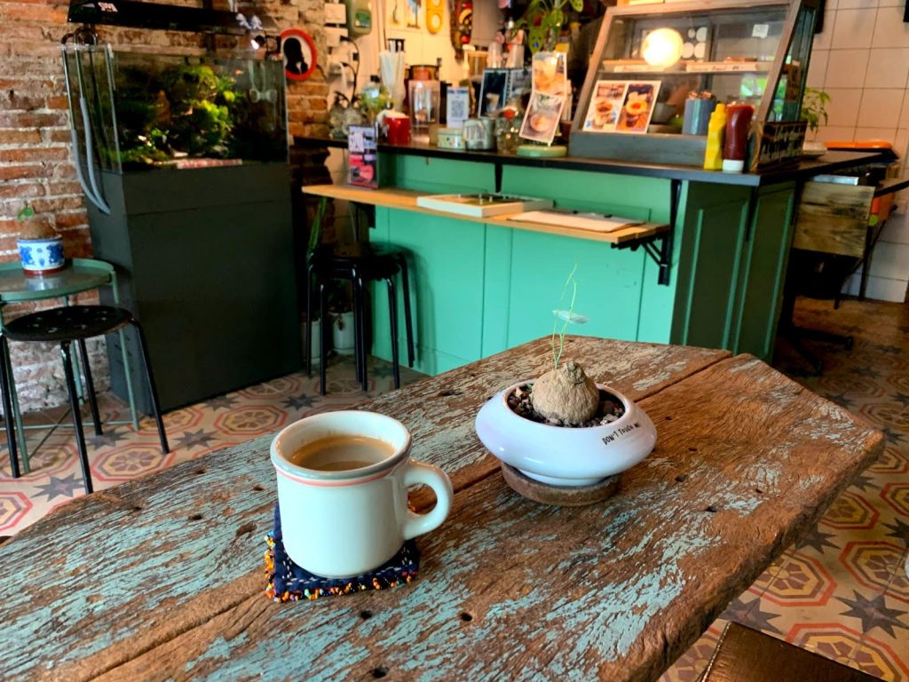 Black coffee in a white ceramic cup on a wooden table inside Suva Cafe, with their green coffee bar in the background.