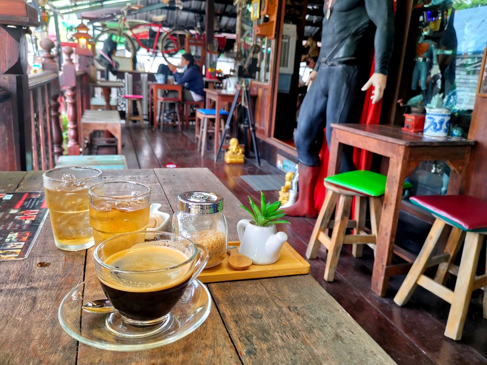 Cup of black coffee beside two glasses of tea and some candies on a table, with colorful movie memorabilia in the background at Toys Cafe Bangkok.