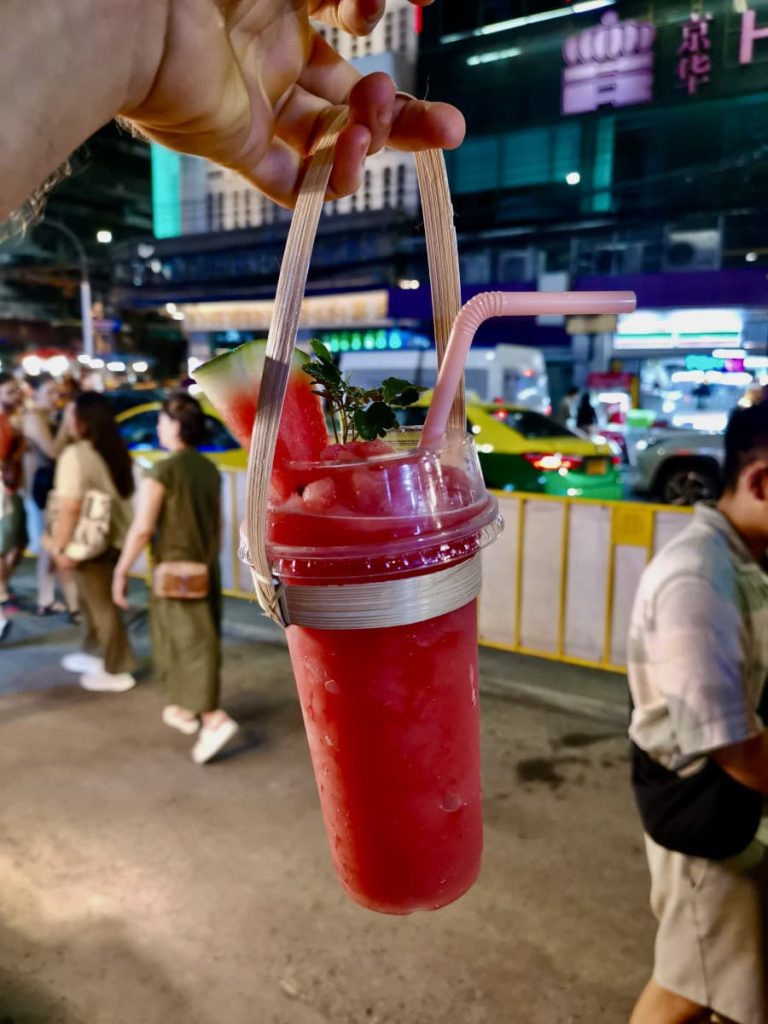 Katharina holding up a plastic cup with thick pink watermelon smoothy inside and a slice of fresh watermelon on top.