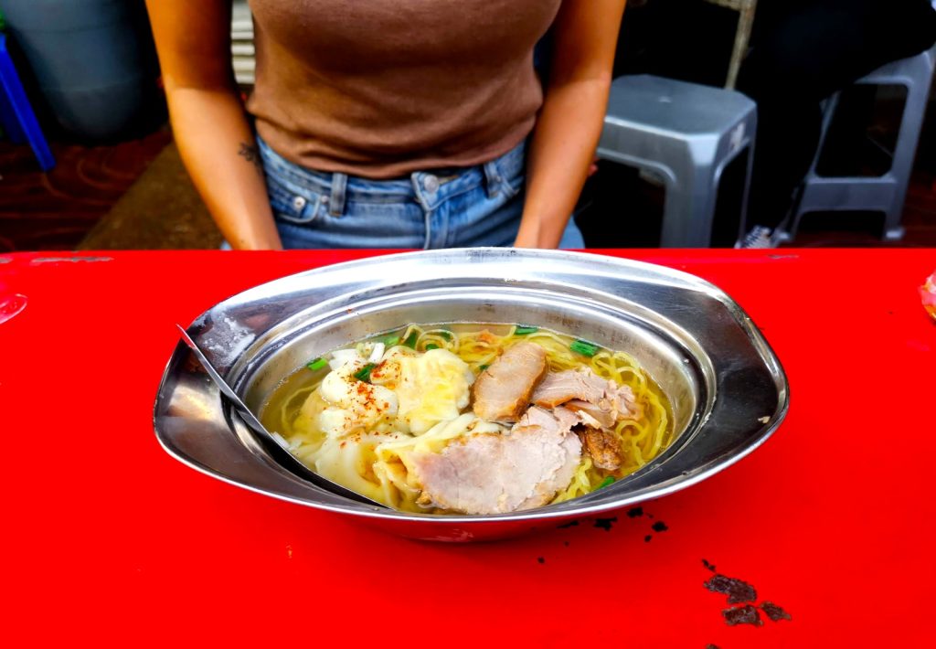 Katharina sitting in front of a red table in Bangkok's Chinatown with a bowl of wonton soup in a metal bowl in front of her.