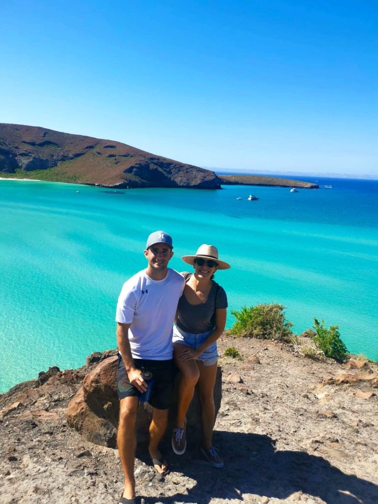 Allan and Katharina (Kat) sit on a rock in front of Balandra Bay in Mexico, with the crystal-clear blue ocean behind them.