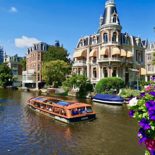 A boat cruising down a canal in amsterdam with some purple flowers in a pot in the foreground