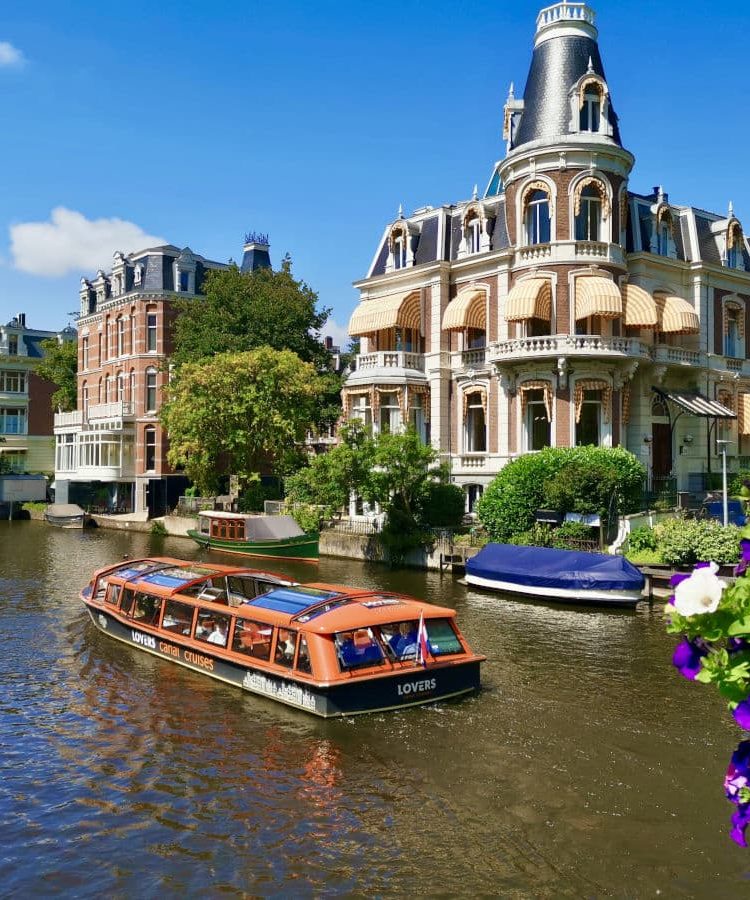 A boat cruising down a canal in amsterdam with some purple flowers in a pot in the foreground