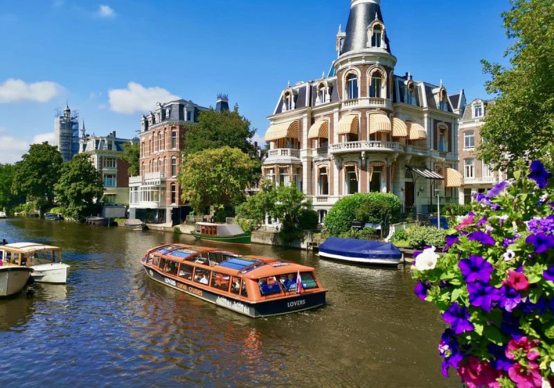 A boat cruising down a canal in amsterdam with some purple flowers in a pot in the foreground