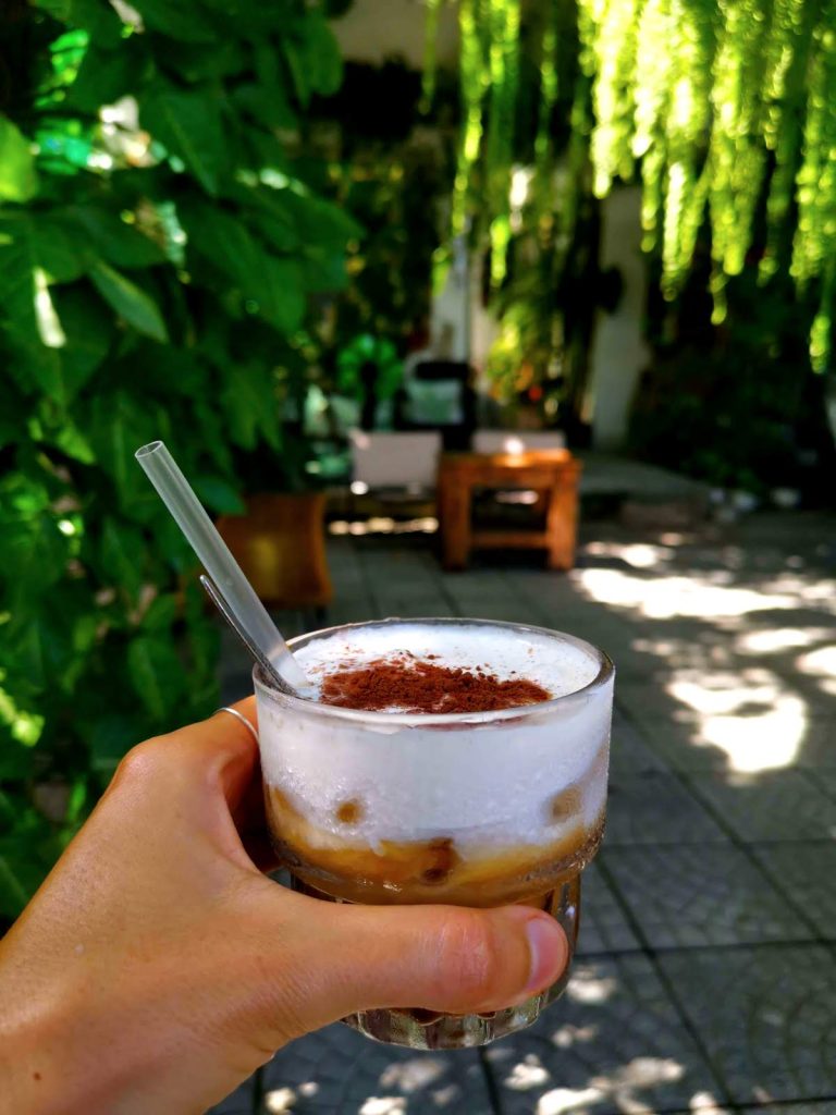 Katharina's hand holding up a small glass with salt coffee at Ba Trong Coffee. In the back are plants and wooden chairs. 