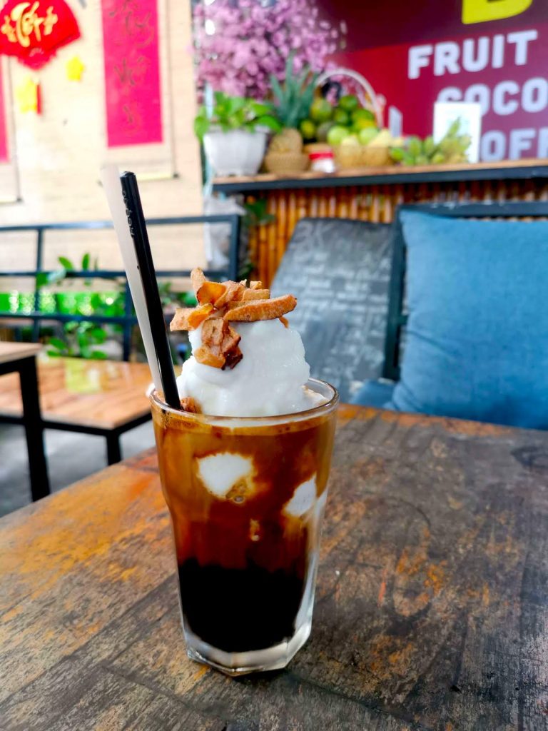 A tall glass with black coffee, coconut cream and roasted coconut flakes on top on a wooden table in front of a sign saying BK Fruit Juice & Coffee Hoi An.