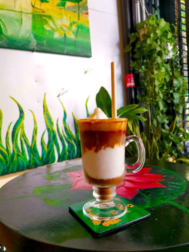 A tall glass with brown coffee, coconut cream and a straw on a table with a painted flower on top and painting in the background at Art Coffee Hoi An.