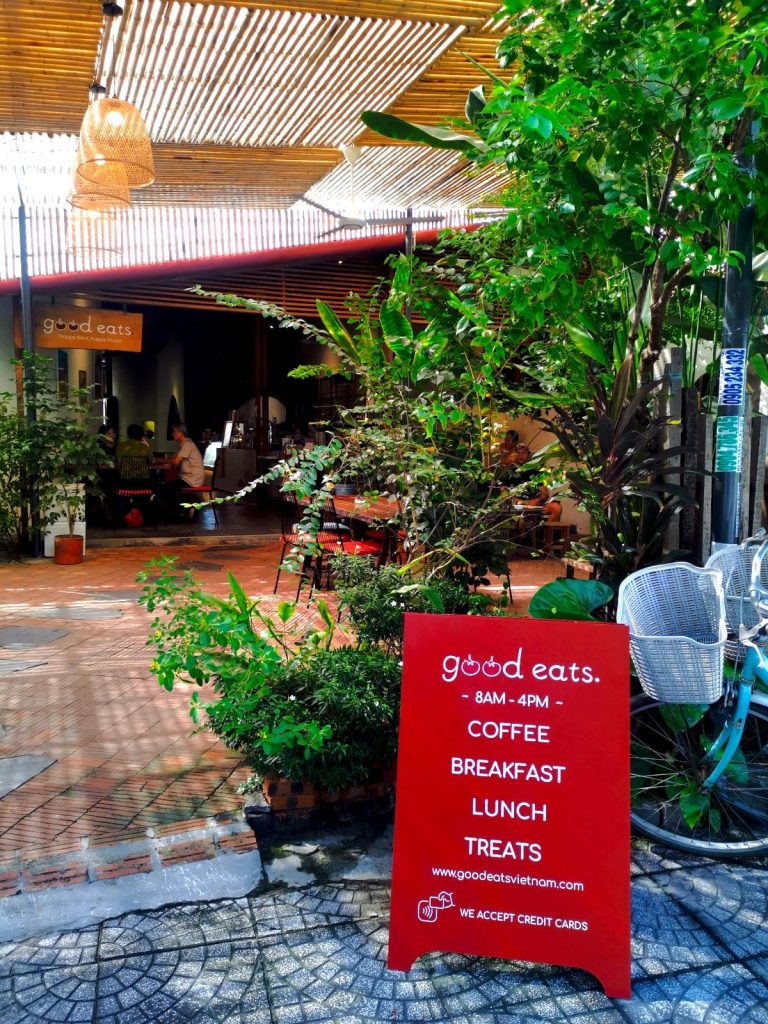 The entrance of Good Eats Hoi An with a red sign advertising coffee, breakfast, lunch and treats, with plants and seats behind.