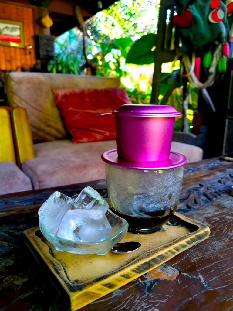 A wooden tray with a bowl of ice cups next to a glass cup with a pink phin filter placed on top at An Nhan Exquisite Cultural Gallery & Coffee on a wooden table in front of a cozy chair.