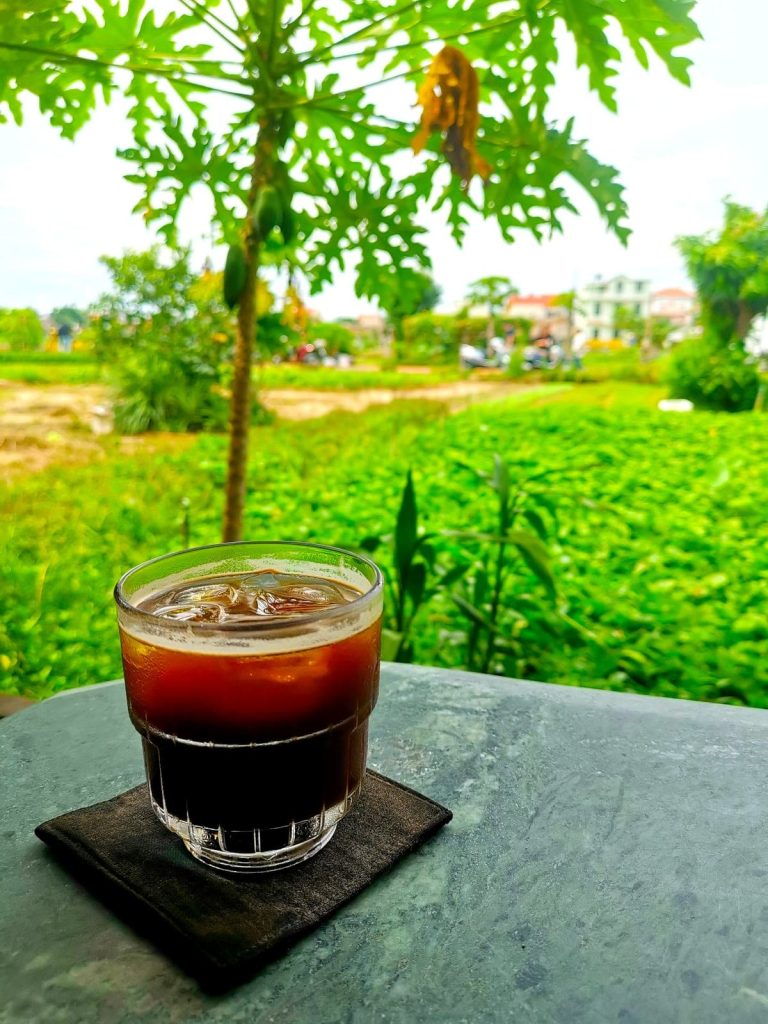 An iced Americano in a glass cup on a table at Cafe Slow Hoi An with a green field in the back.