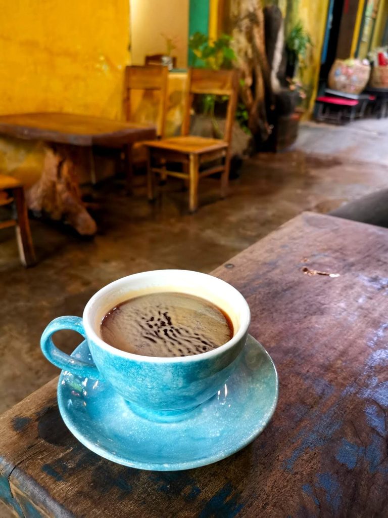 A hot Americano in a blue ceramic cup on a wooden table at Hoi An Coffee Hub.