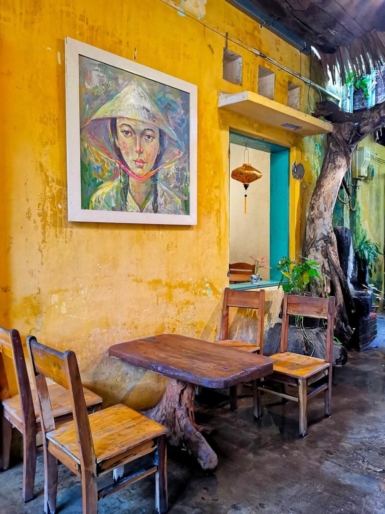 Four wooden chairs around a wooden table in front of a yellow wall with a painting on it at Hoi An Coffee Hub.