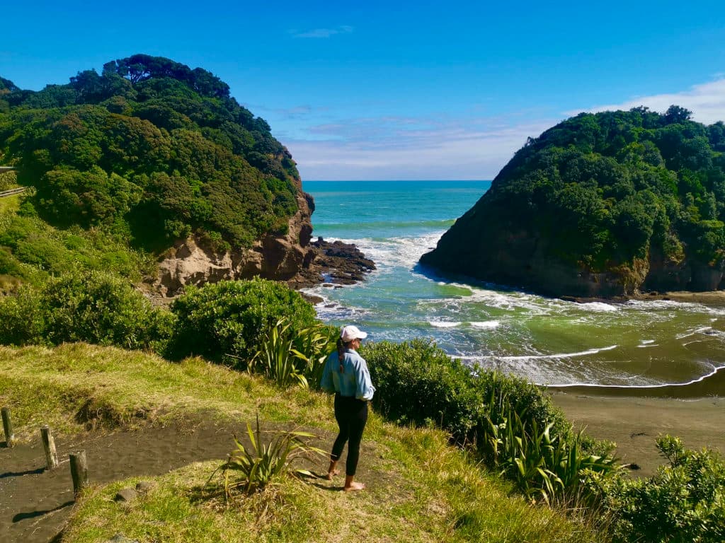 Katharina overlooks the rocky, green bay and blue ocean at Bethells Beach in Auckland, New Zealand.