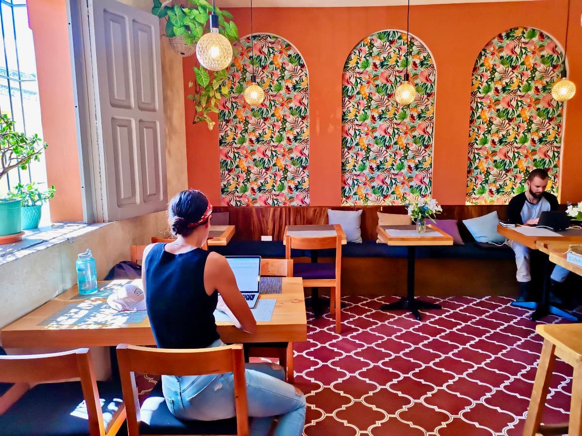 Katharina, working remotely as a digital nomad, sits in a cafe with bright orange walls and a red floor, using her laptop.