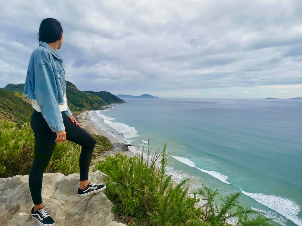 Katharina, in a blue denim jacket, overlooks the blue-green ocean in front of Mangawhai Hill, New Zealand.