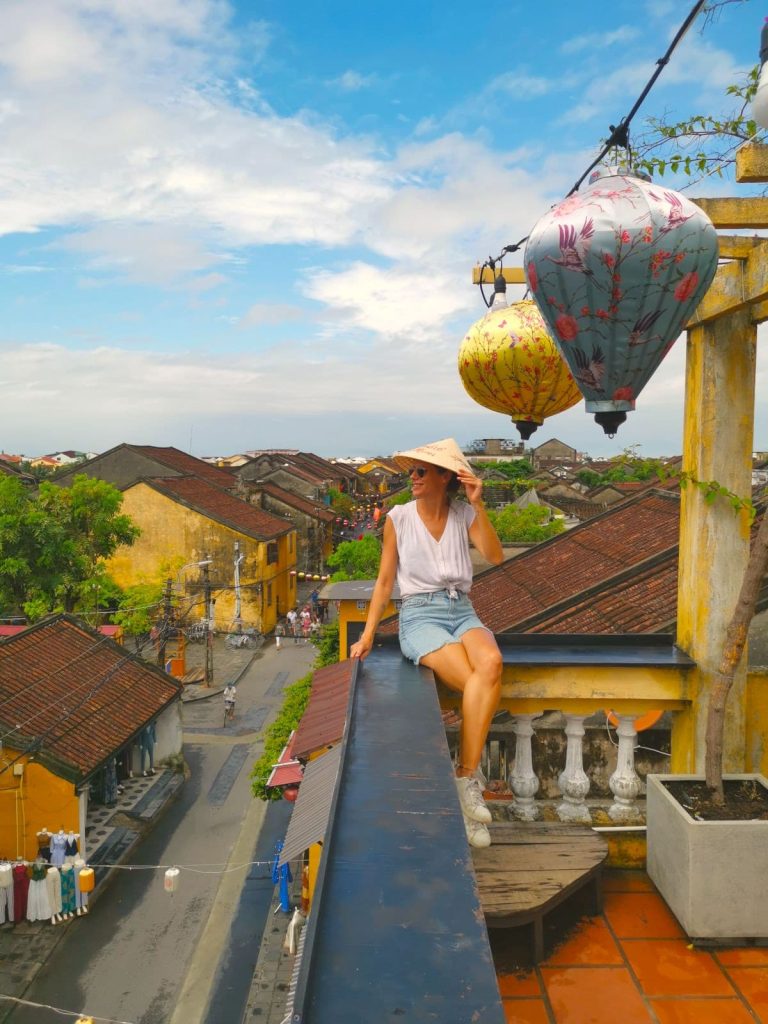 Katharina wearing a white shirt and blue shorts, holding up a traditional Vietnamese conical hat, seated on the edge of Memories Cafe rooftop with a view of Hoi An's streets below.