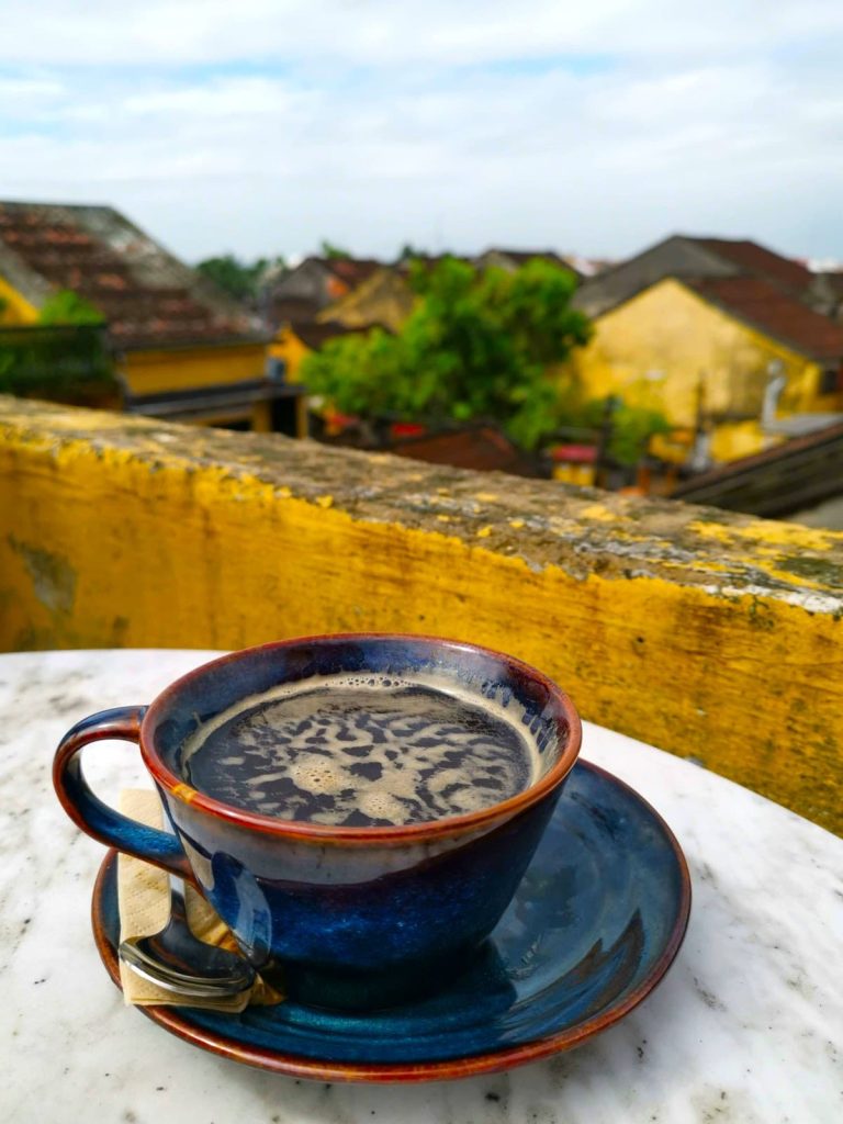 A hot Americano in a dark blue ceramic cup sitting on a white marble table with yellow buildings in the background at Memories Rooftop Brunch & Cafe Hoi An.