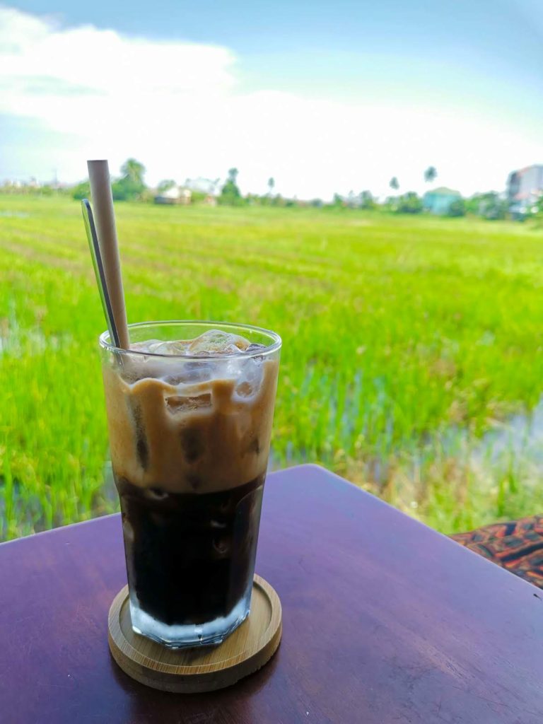 A tall glass cup with ice and black coffee and a straw on a wooden table at NGHE Rice Paddy Cafe Hoi An with a green rice field in the background.