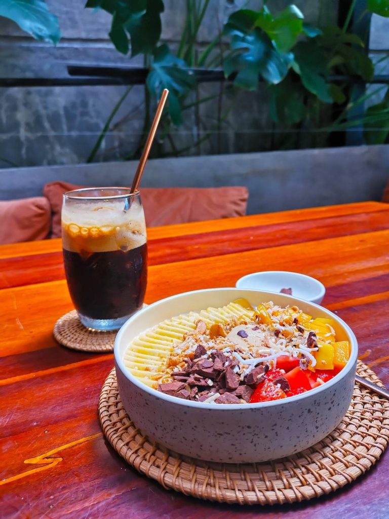 A glass with iced black coffee on a table next to a white bowl filled with fruits and granola at Nourish Eatery Hoi An.