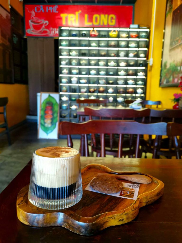 A wooden tray with a glass filled with black coffee and a creamy foam next to a cookie at Tri Long Cafe Hoi An. 