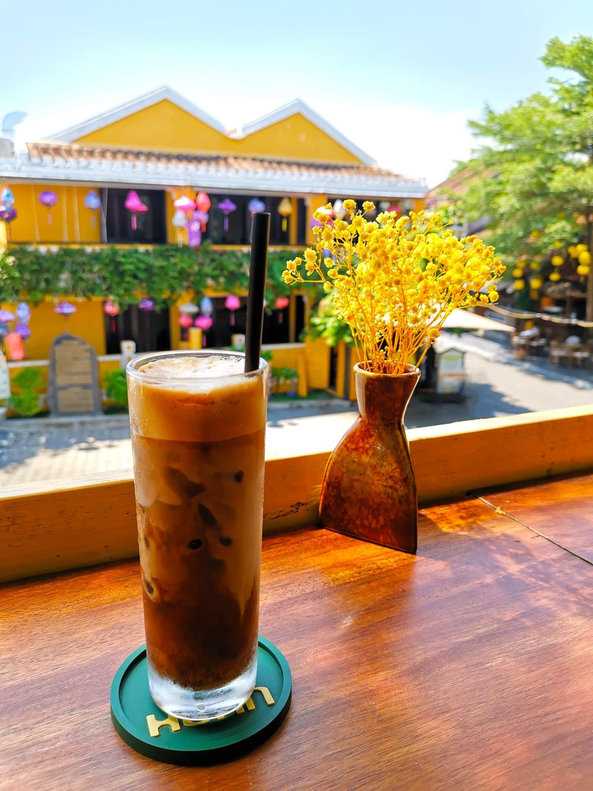 A tall glass of Vietnamese black coffee with milk foam on top on a wooden bar at Uncle Huan Cafe, overlooking the streets of the Ancient Town of Hoi An.