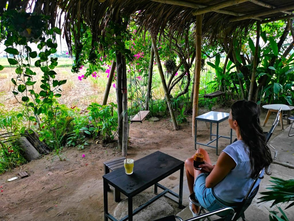 Katharina sitting on a small chair with a glass of black coffee in her hands at Xom Chieu Cafe, looking out into a rice field with plants and pink flowers around it.